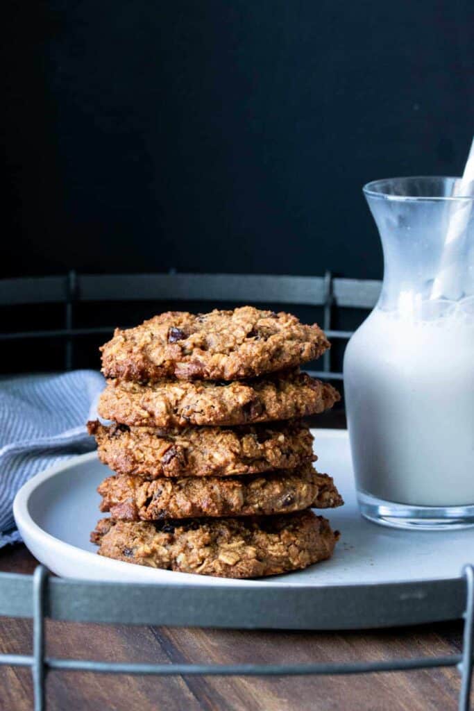 Vegan gluten free oatmeal raisin cookies stacked on a white plate next to a pitcher of milk.