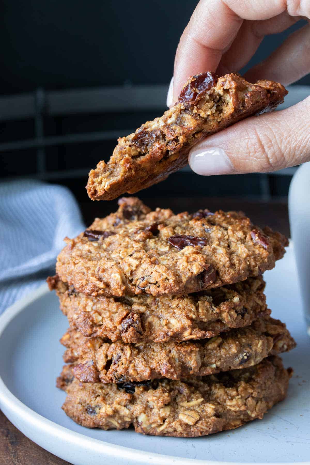 Hand holding half eaten gluten free oatmeal raisin cookie with a stack of cookies on white plate.