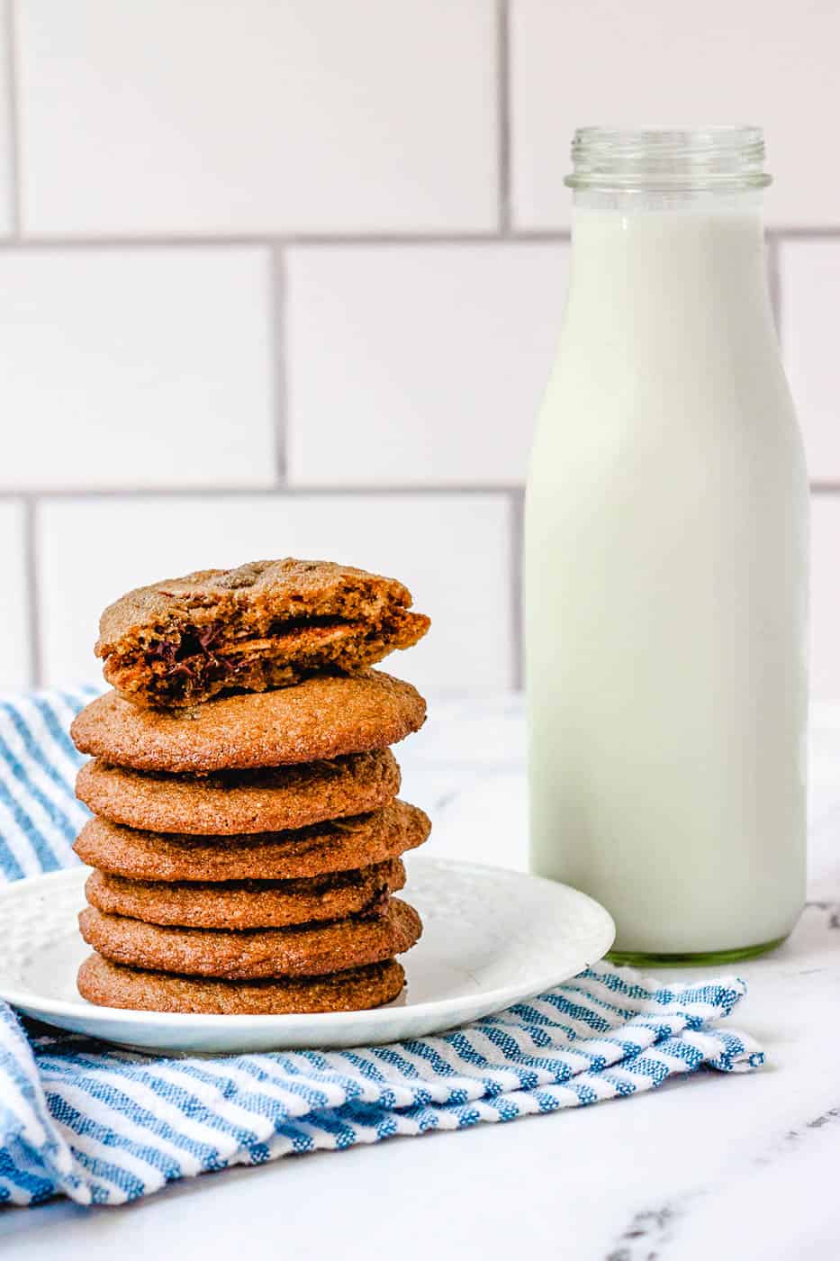 healthy chocolate chip cookies stacked on a cutting board, with one cookie broken for a closeup inside and milk in the background