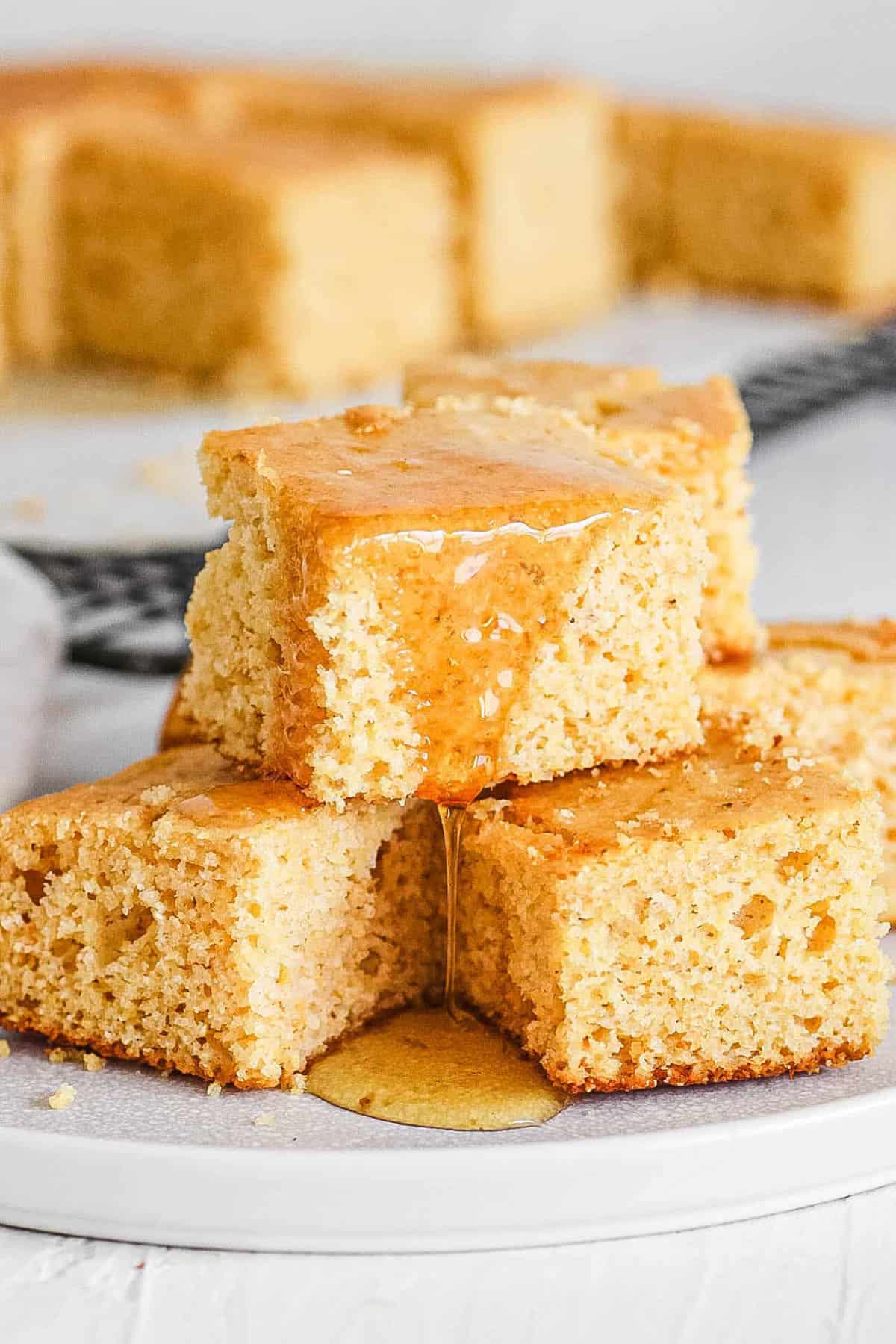 Healthy cornbread squares drizzled with honey, stacked on a white plate.