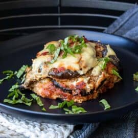 Black plate with a piece of vegan gluten-free eggplant parmesan topped with sliced basil.