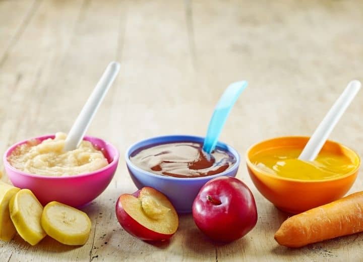 pureed food in small serving bowls including pureed bananas in pink bow, pureed plums in purple bowl, and pureed carrots in orange bowl