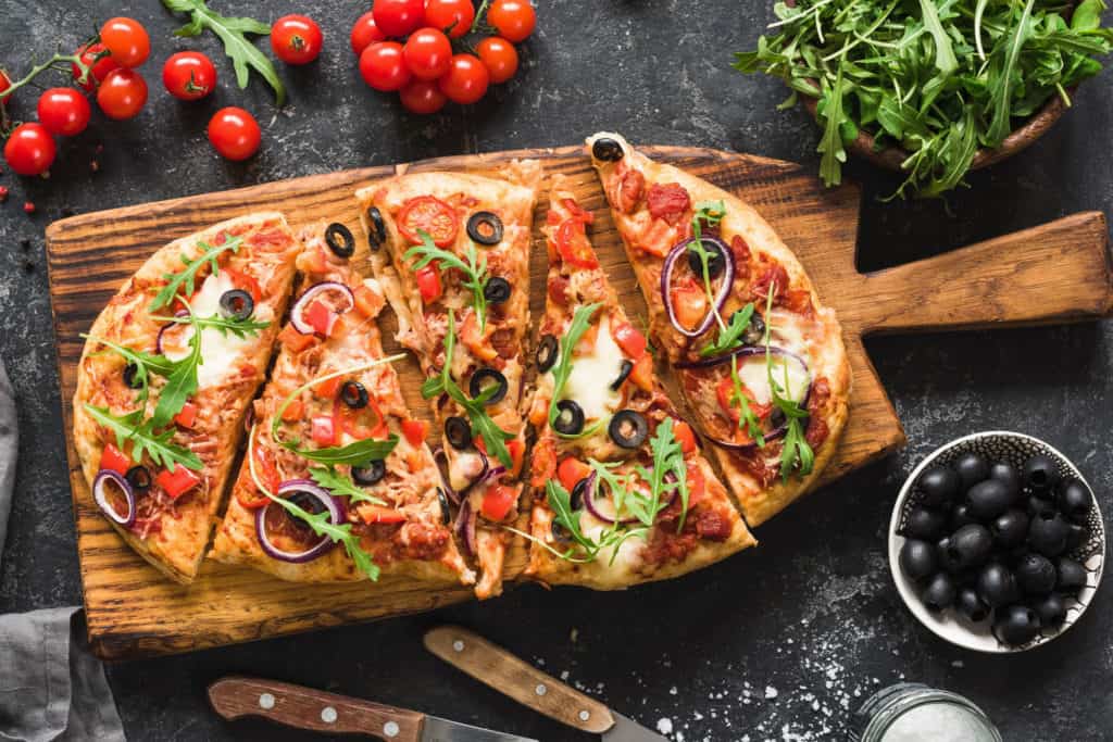 Flatbread pizza garnished with fresh arugula on wooden pizza board, top view. Dark stone background - healthy pizza toppings