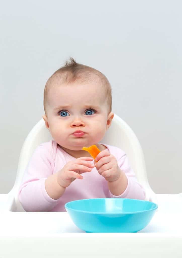 baby in high chair with blue bowl in front of her holding butternut squash bites