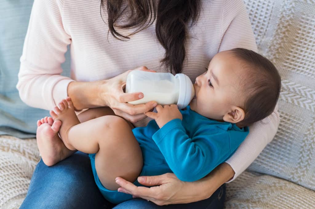 probiotics for baby constipation - Little infant baby lying on mothers hand drinking milk from bottle. Newborn baby drinking milk from bottle. Cute toddler with milk bottle on leg of mother.