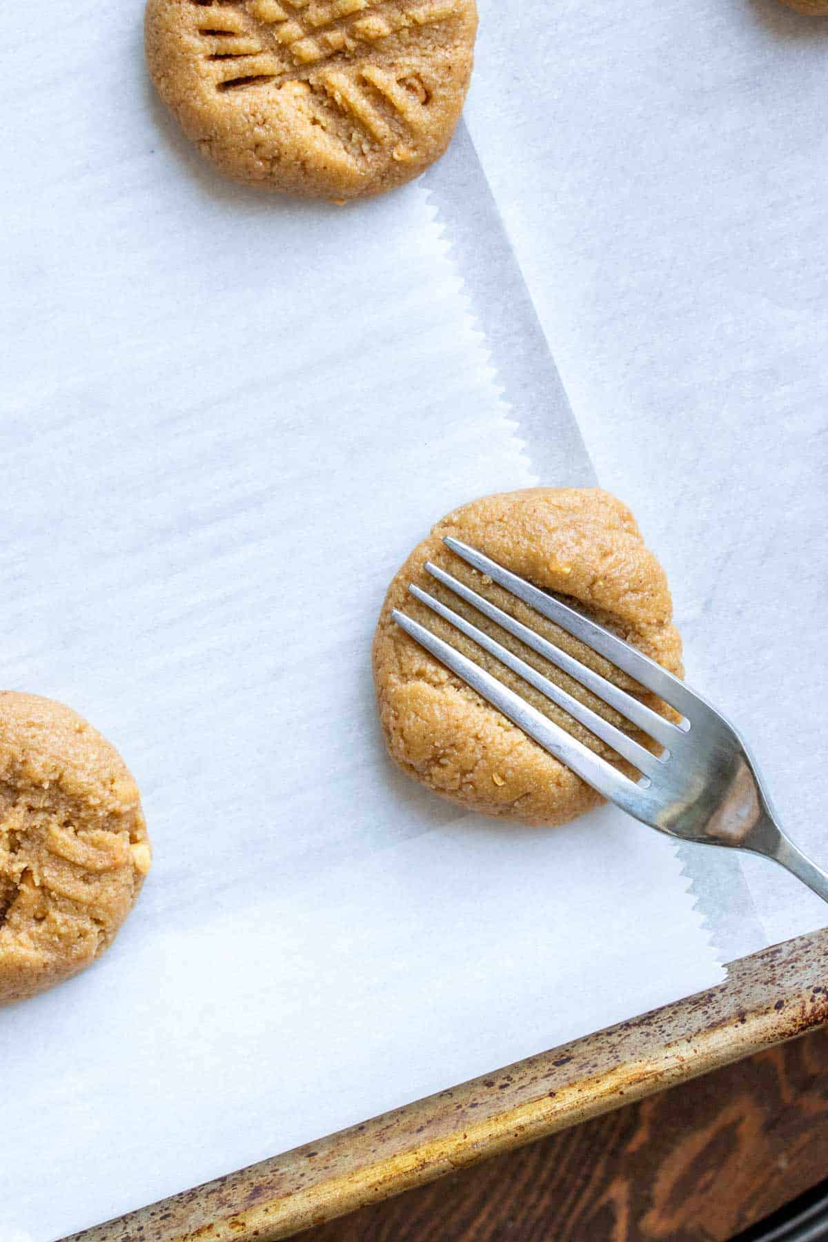 Peanut butter cookies on parchment paper, with a fork pressing a design into one of the cookies.