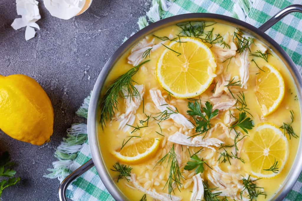 avgolemono - delicious creamy greek chicken soup with lemon, egg yolk, rice and herbs in a casserole on a concrete table with ingredients at the background, flatlay, close-up