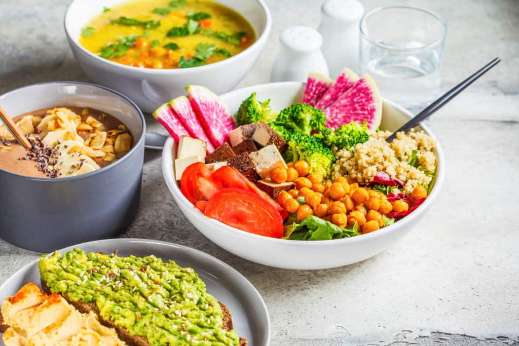 vegan lunch ideas - Vegan lunch table. Chocolate smoothie bowl, Buddha bowl with tofu, chickpeas and quinoa, lentil soup and toasts on a gray background.