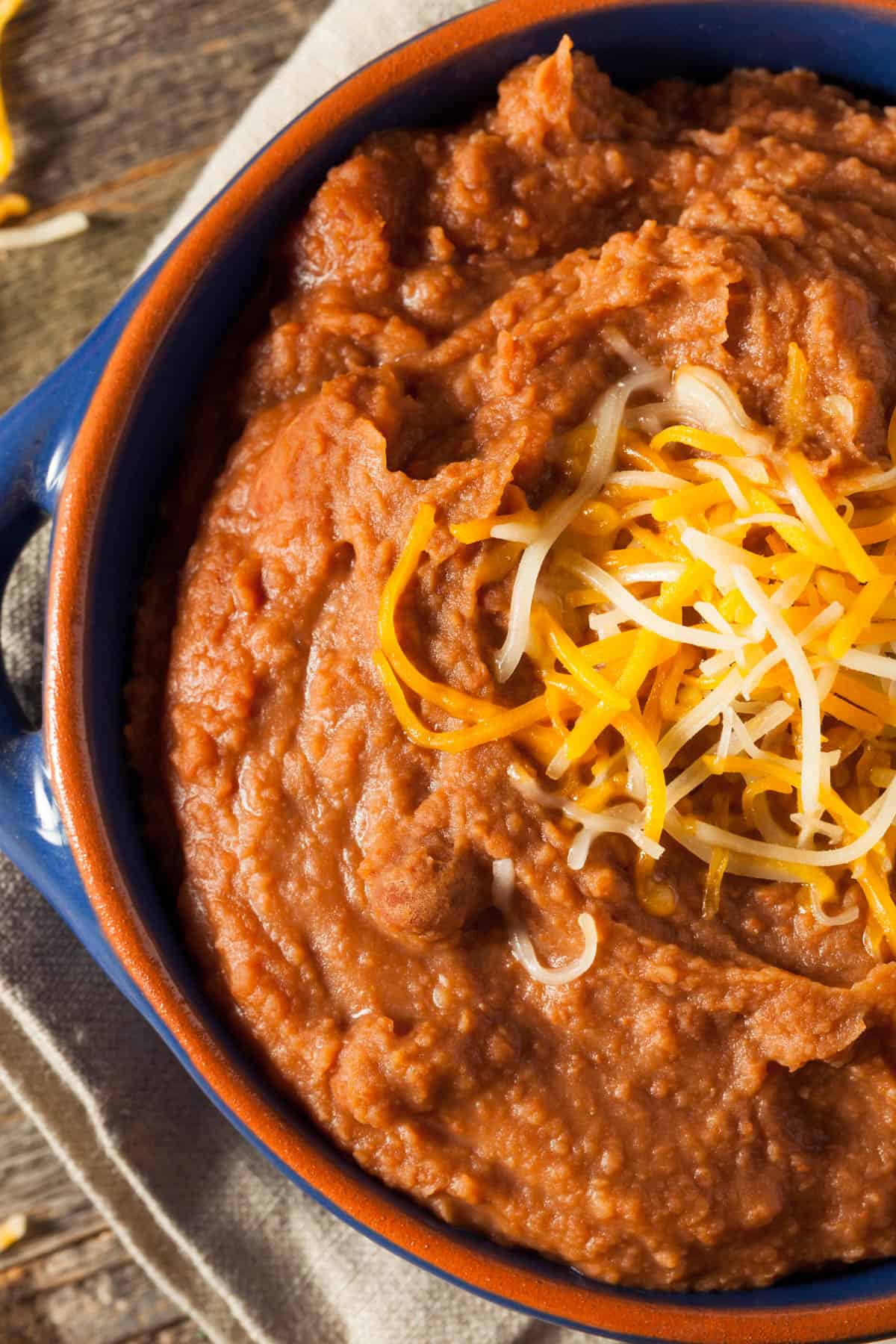 Blue bowl filled with vegan refried beans topped with vegan shredded cheese on a tan towel.