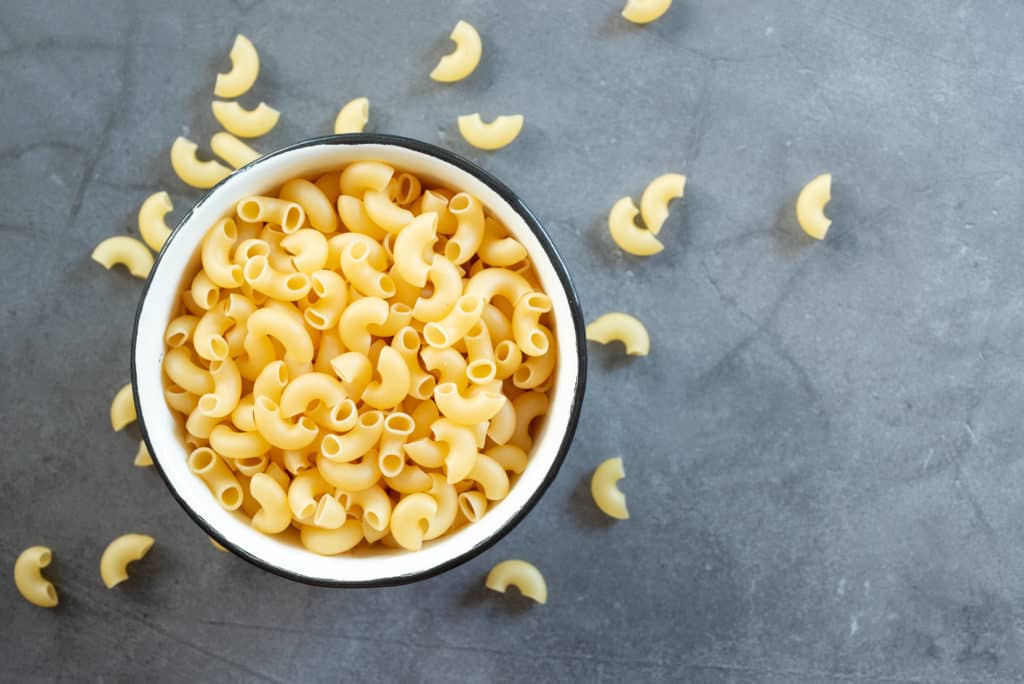 foods that s، with e - raw elbow macaroni in enamel bowl on dark cement table background. 