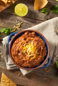 Blue bowl filled with vegan refried beans topped with vegan shredded cheese on a tan towel.