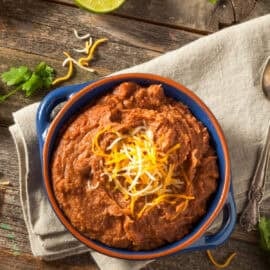 Blue bowl filled with vegan refried beans topped with vegan shredded cheese on a tan towel.