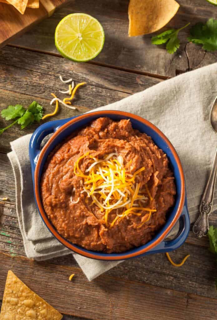Blue bowl filled with vegan refried beans topped with vegan shredded cheese on a tan towel.