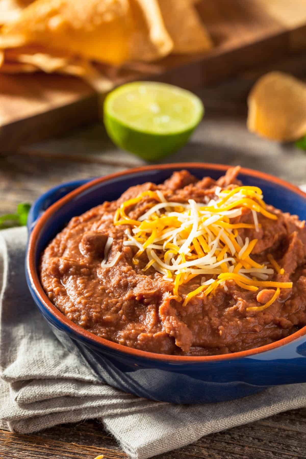 Blue bowl filled with vegan refried beans topped with vegan shredded cheese on a tan towel.