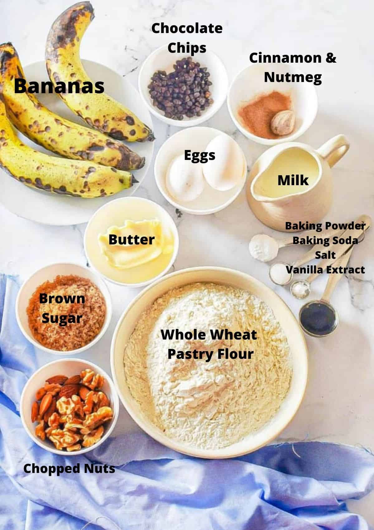 Ingredients for chocolate chip banana muffins on counter top: bananas, eggs, butter, spices, milk, baking powder, baking soda, salt, vanilla, whole wheat pastry flour, chopped nuts, and brown sugar.