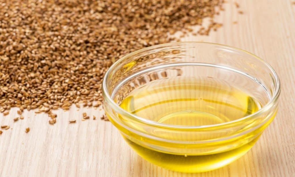 Flaxseed oil in small glass bowl beside flaxseeds on wood countertop.