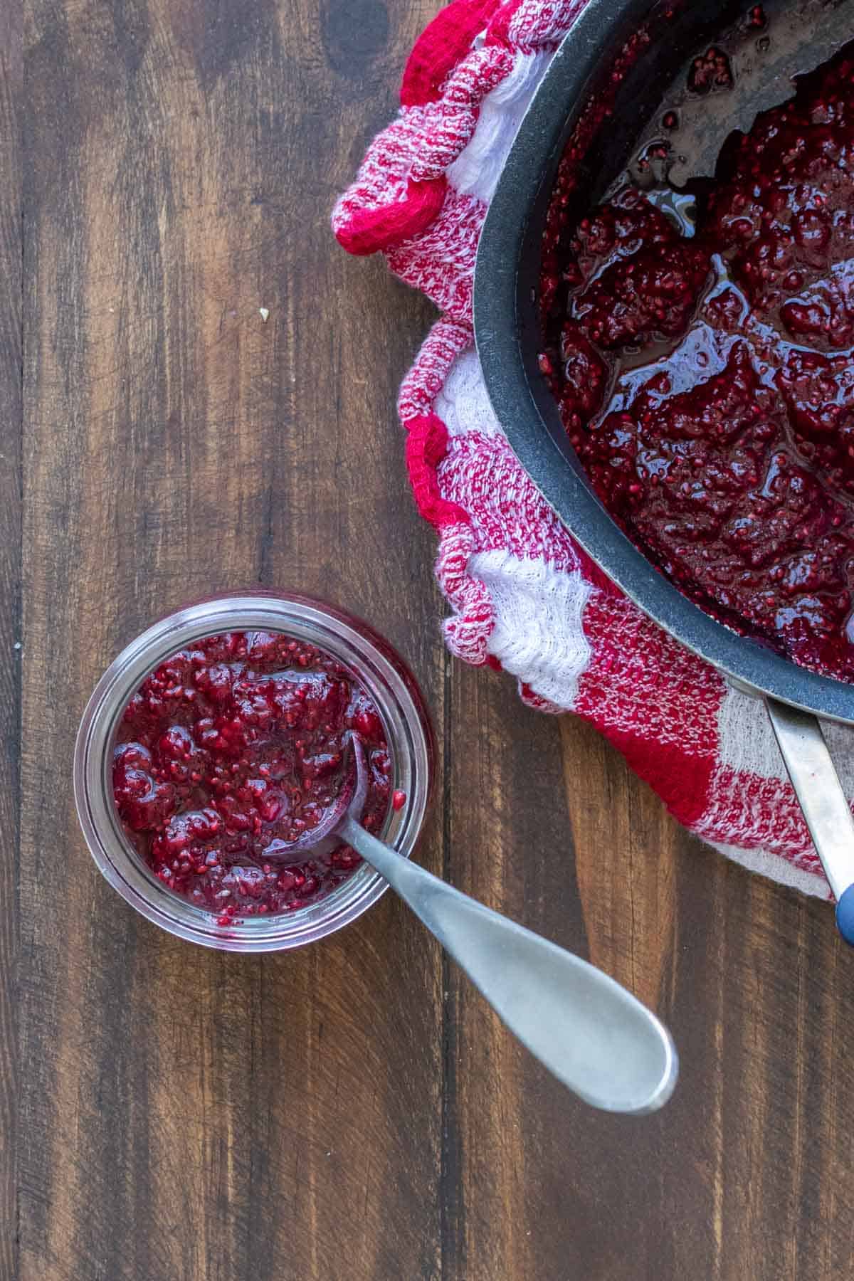 Overhead shot of strawberry chia seed jam in a glass jar with spoon, with pot of jam on the side.