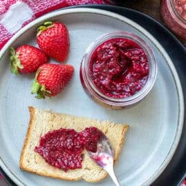 Strawberry chia seed jam, in a mason jar on a plate next to a piece of toast with jam spread on it.