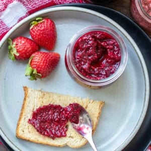 Strawberry chia seed jam, in a mason jar on a plate next to a piece of toast with jam spread on it.