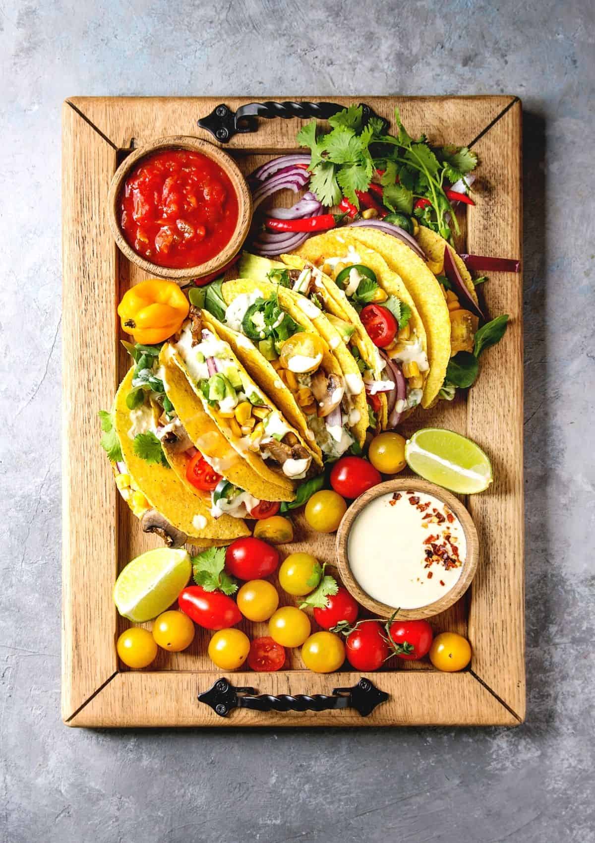 Vegetarian tacos with lots of taco toppings on wood cutting board, surrounded by taco toppings such as salsa, cilantro, onion, peppers, tomatoes, and a creamy dressing. 