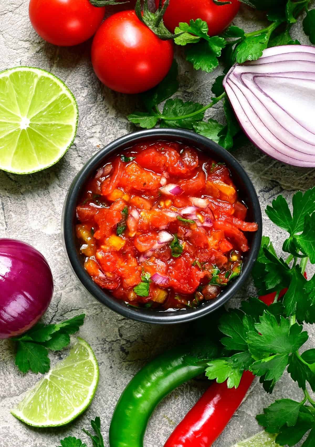Tomato salsa in small black bowl, surrounded by ingredients such as red onion, lime, jalapeño pepper.