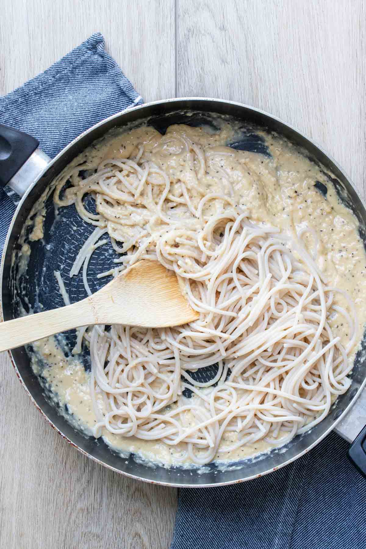 Wooden spoon mixing spaghetti into a creamy sauce in a saute pan.