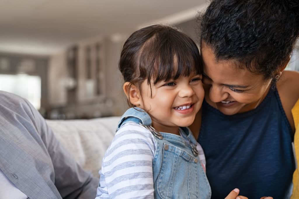 Beautiful black mother embracing little girl sitting on couch with copy space. Cute daughter hugging african american mother and smiling together. Happy latin mom jokes with her kid at home and playing together