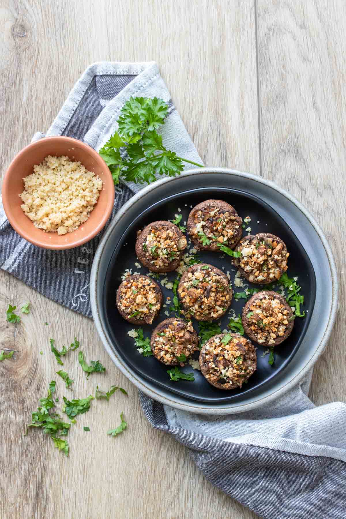 Top view of a plate of stuffed appetizer mushrooms topped with parsley next to a peach bowl of Parmesan