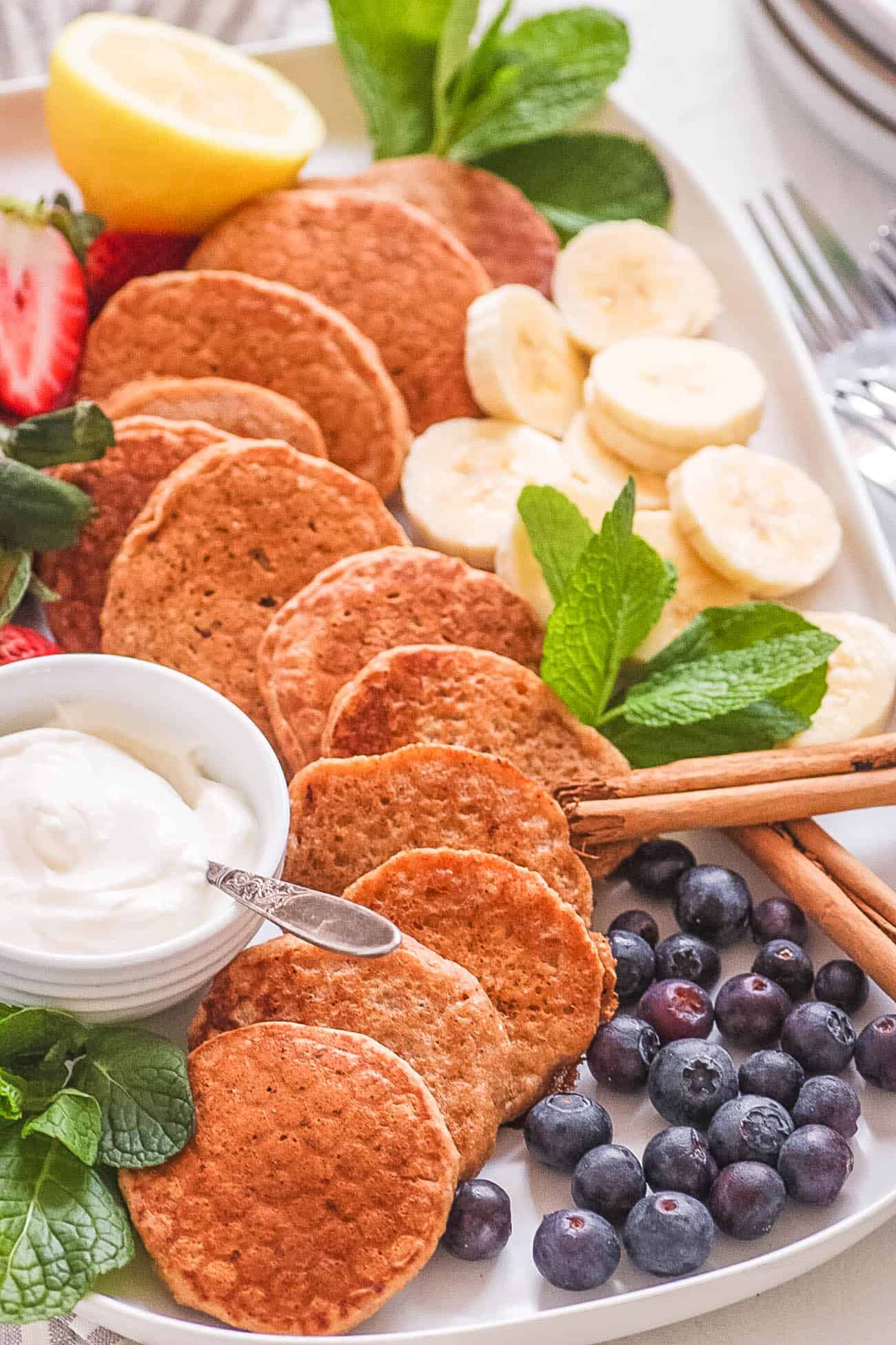 A white platter of mini pancakes with fruit, yogurt, and cinnamon sticks.