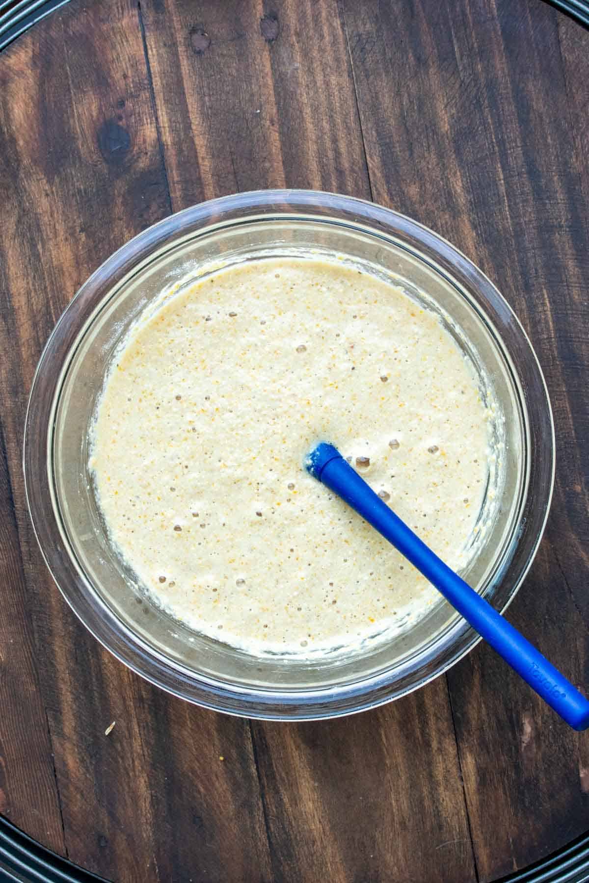 Blue spatula mixing cornbread batter in a glass bowl.