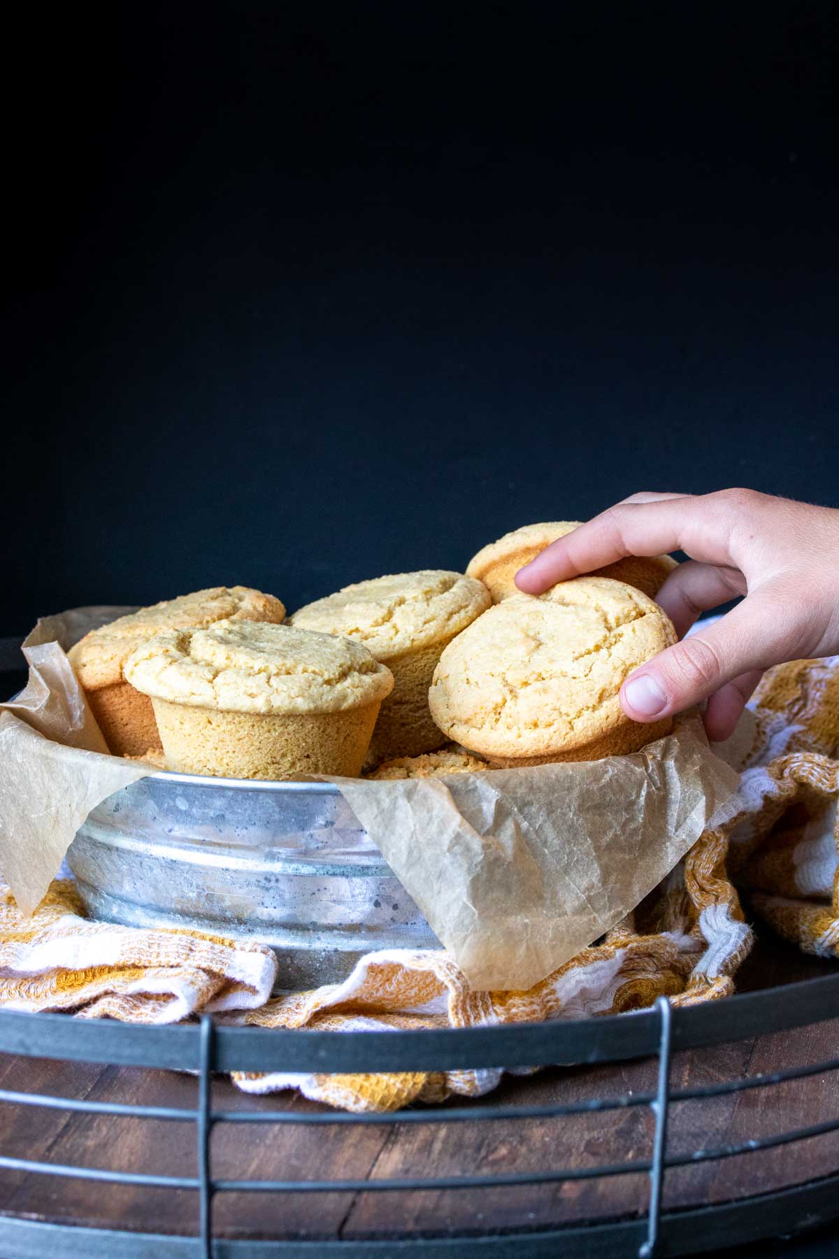 Hand grabbing a vegan cornbread muffin from a metal bowl on a yellow checkered towel.