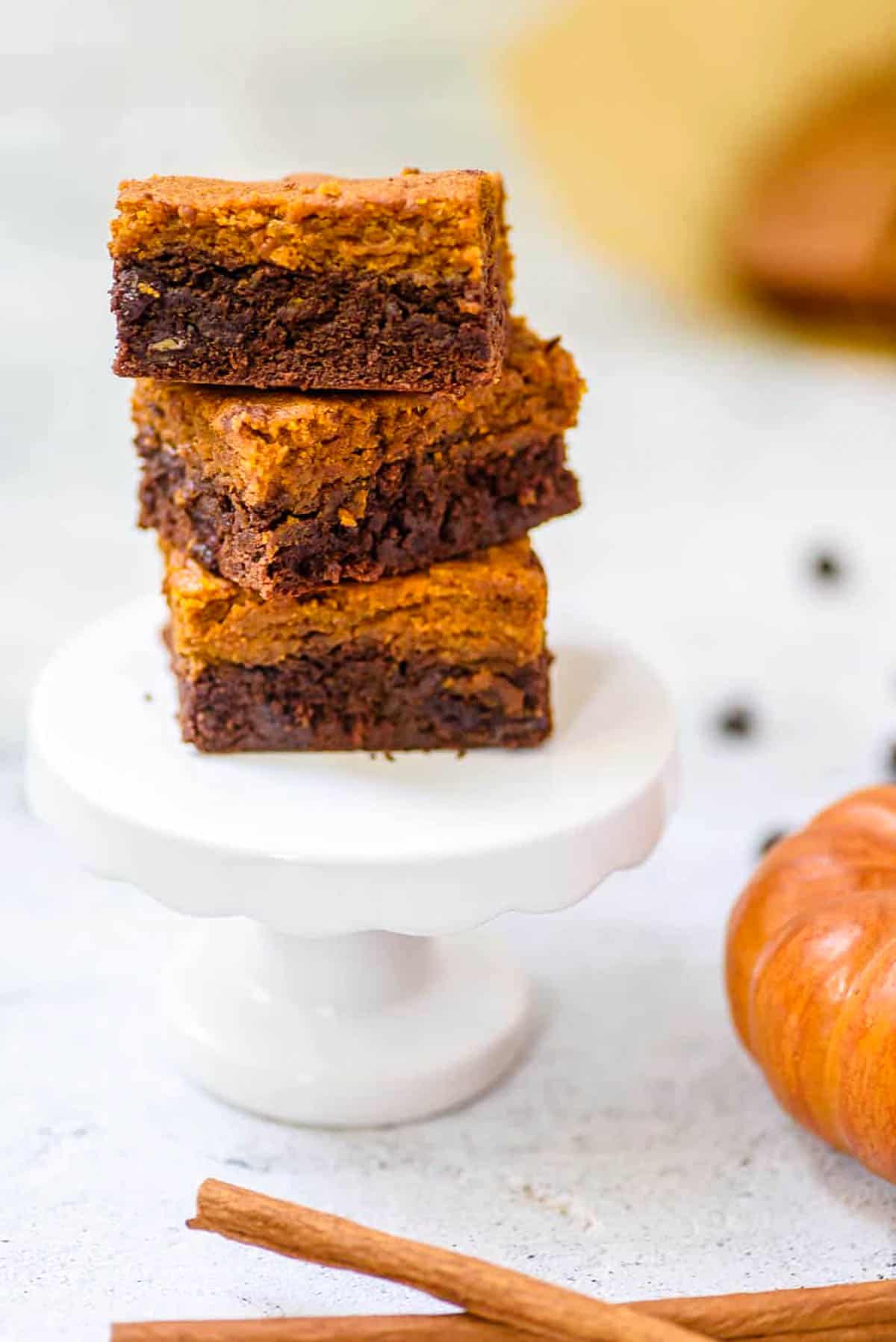 Healthy pumpkin brownies stacked on a white pedestal, with cinnamon, chocolate chips and pumpkin in the background.