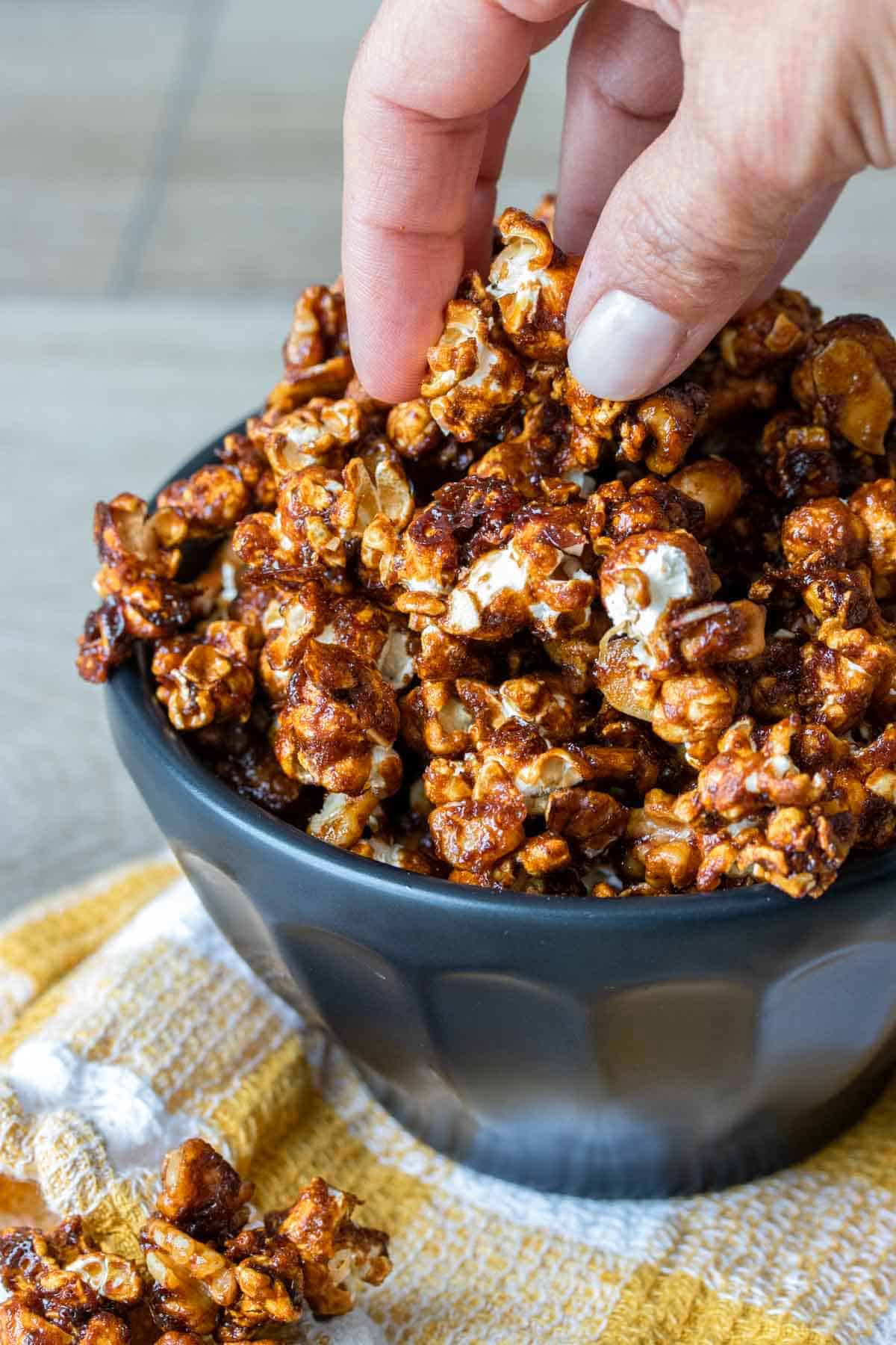 Someone's hand reaching for a piece of vegan caramel popcorn in a bowl.