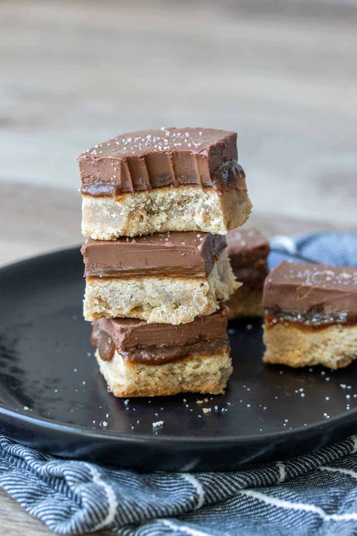 A stack of vegan millionaire shortbread squares on a black plate with the top one half eaten.