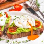 Two slices of avocado toast with poached eggs and tomato served on a white plate.