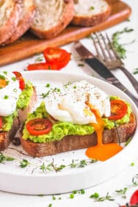 Two slices of avocado toast with poached eggs and tomato served on a white plate.