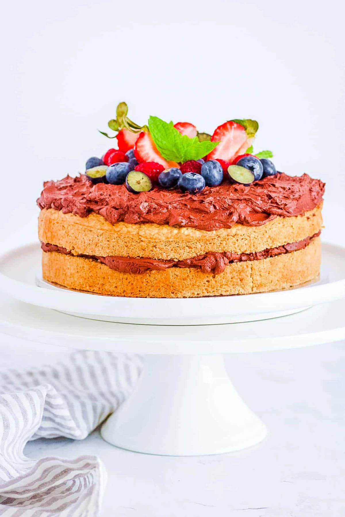 Fluffy vegan oat flour cake topped with berries and mint, on a cake stand.