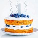 A side shot of a healthy smash cake on a white plate. The cake is topped with blueberries, a number 1 candle, and silver candles.