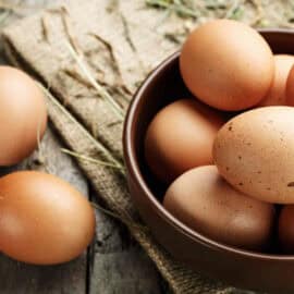 Farm fresh eggs stored in a grey bowl on a brown countertop with a couple of fresh eggs outside of the bowl too.