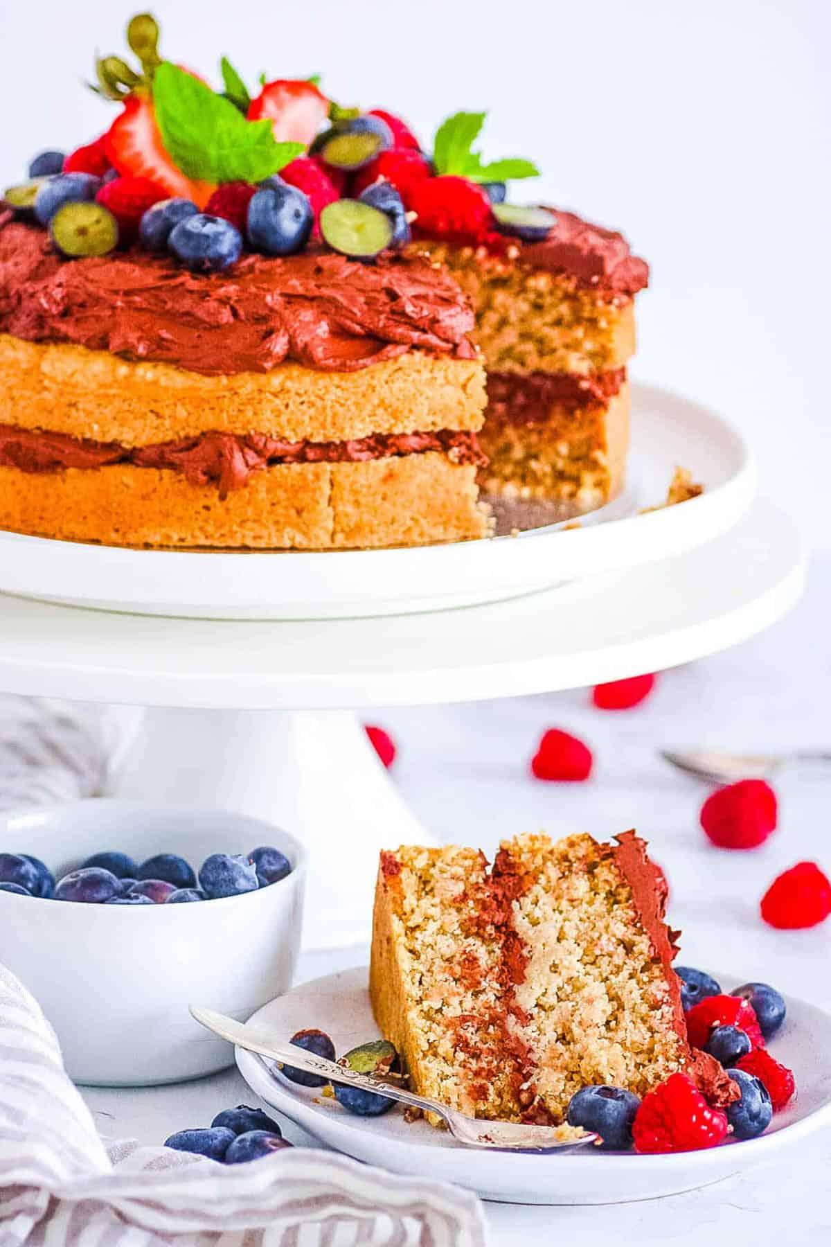 Vanilla vegan oat flour cake topped with berries and mint, on a cake stand with a slice of cake on a plate next to it.