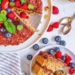 Gluten free, vegan oat flour cake topped with berries and mint, on a cake stand with a slice of cake on a plate next to it.