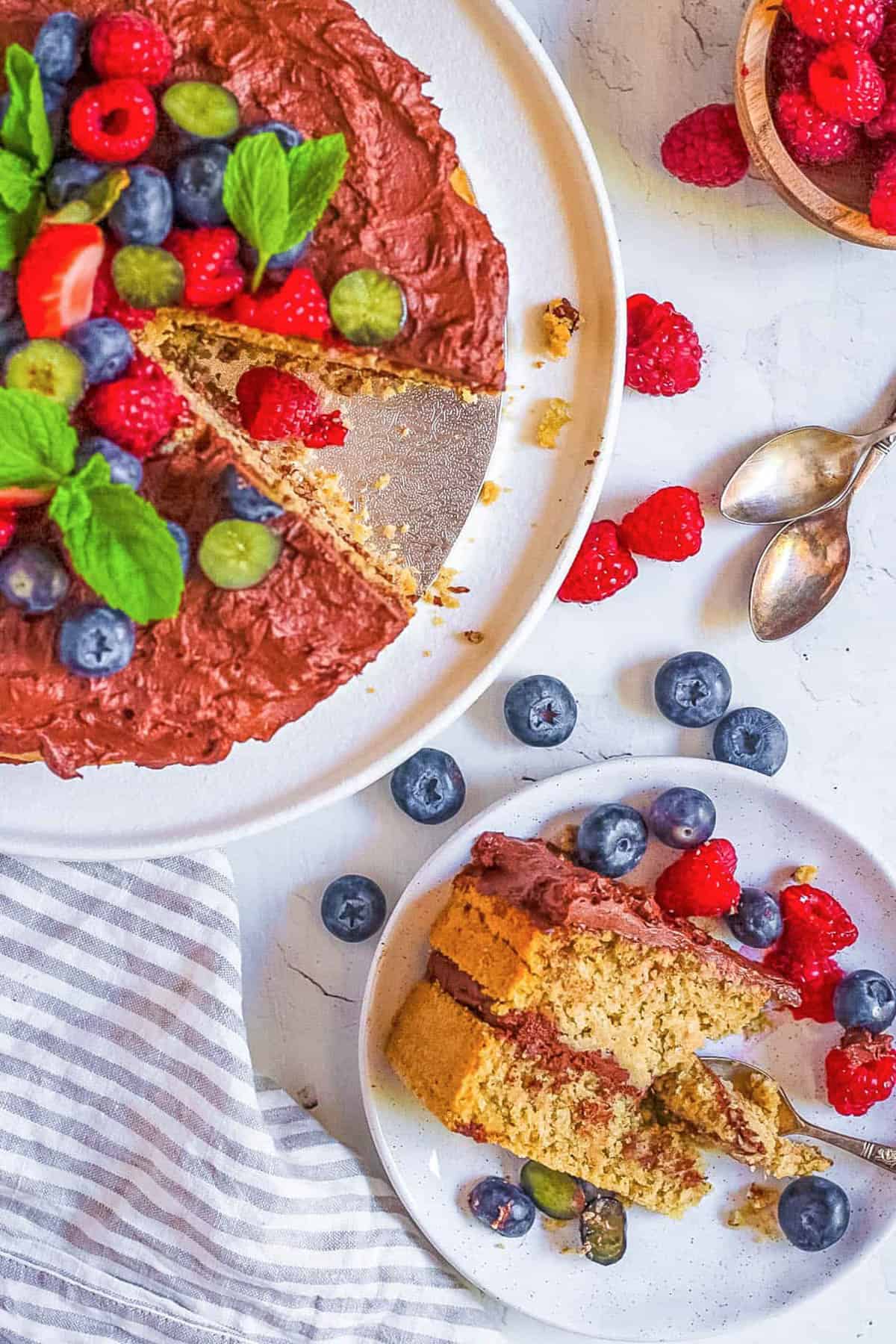 Gluten free, vegan oat flour cake topped with berries and mint, on a cake stand with a slice of cake on a plate next to it.