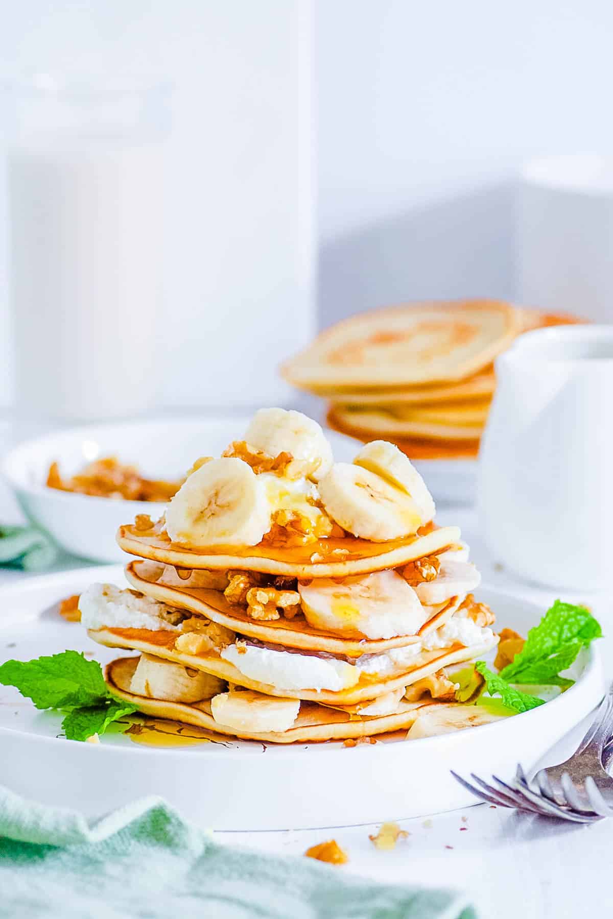 A stack of protein powder pancakes on a white plate with whipped cream, bananas, and walnuts.