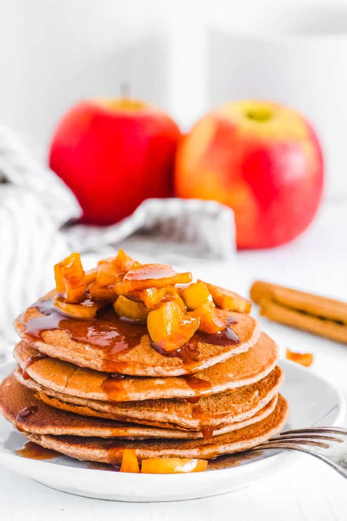 A stack of apple cinnamon pancakes on the counter with apple compote and maple syrup on top.