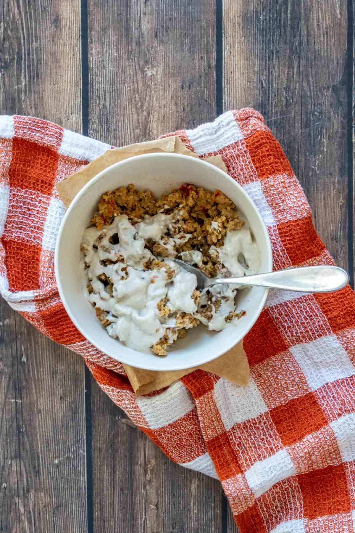 A spoon mixing crumbled sausage and a creamy sauce in a cream colored bowl on a dark orange checkered towel.