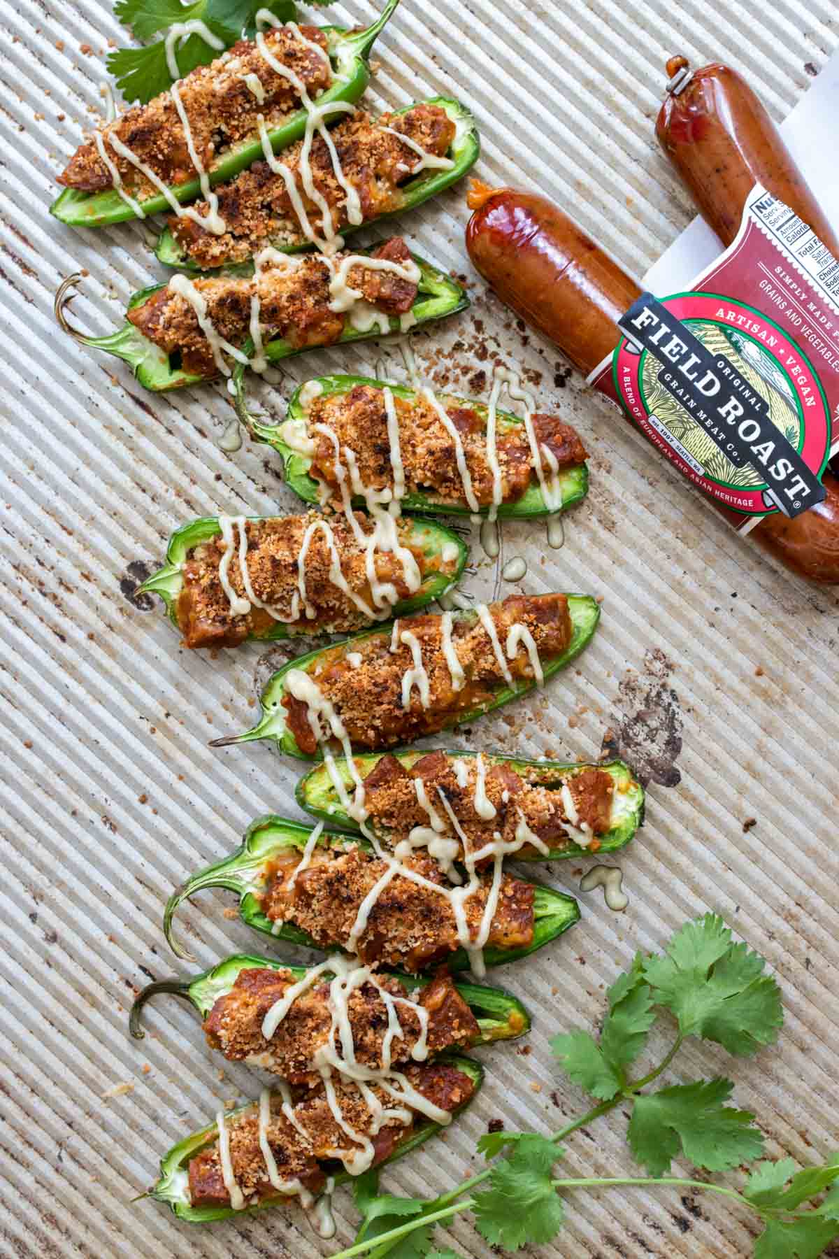 Baking sheet with vegan stuffed jalapeños drizzled with cheese sauce next to package of sausages.