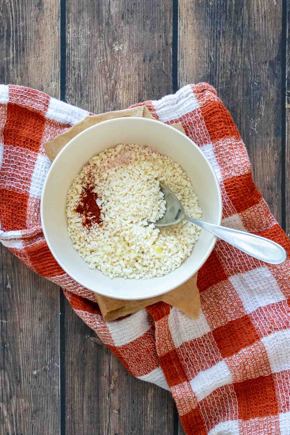 A cream bowl with a spoon in it and breadcrumbs and spices on a dark orange checkered towel.