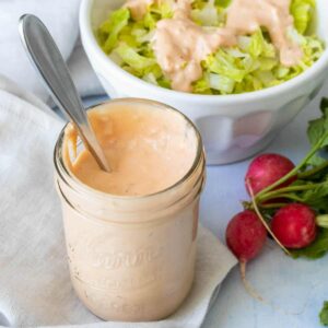 A glass jar with dairy-free thousand island dressing sitting on a light colored napkin in front of a bowl of salad.