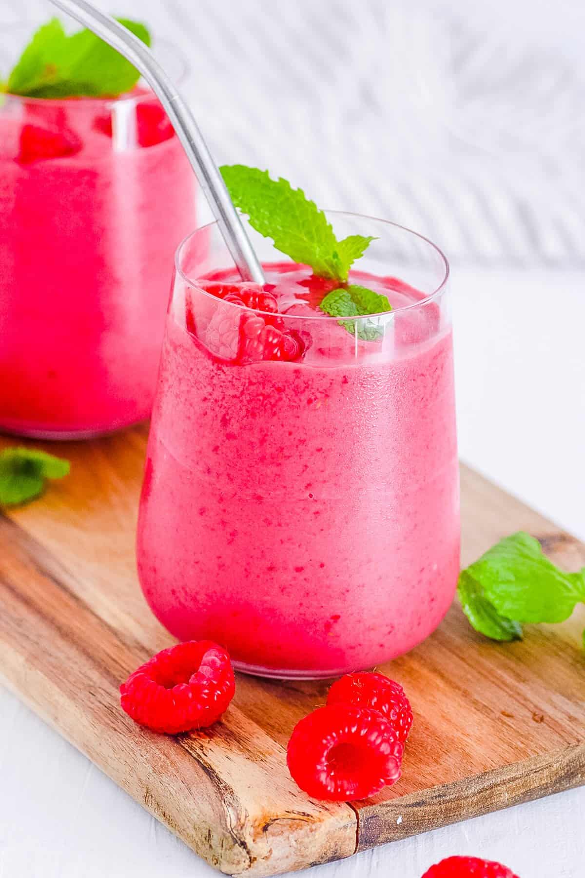 Two glasses of raspberry smoothie with mint on a wooden cutting board.
