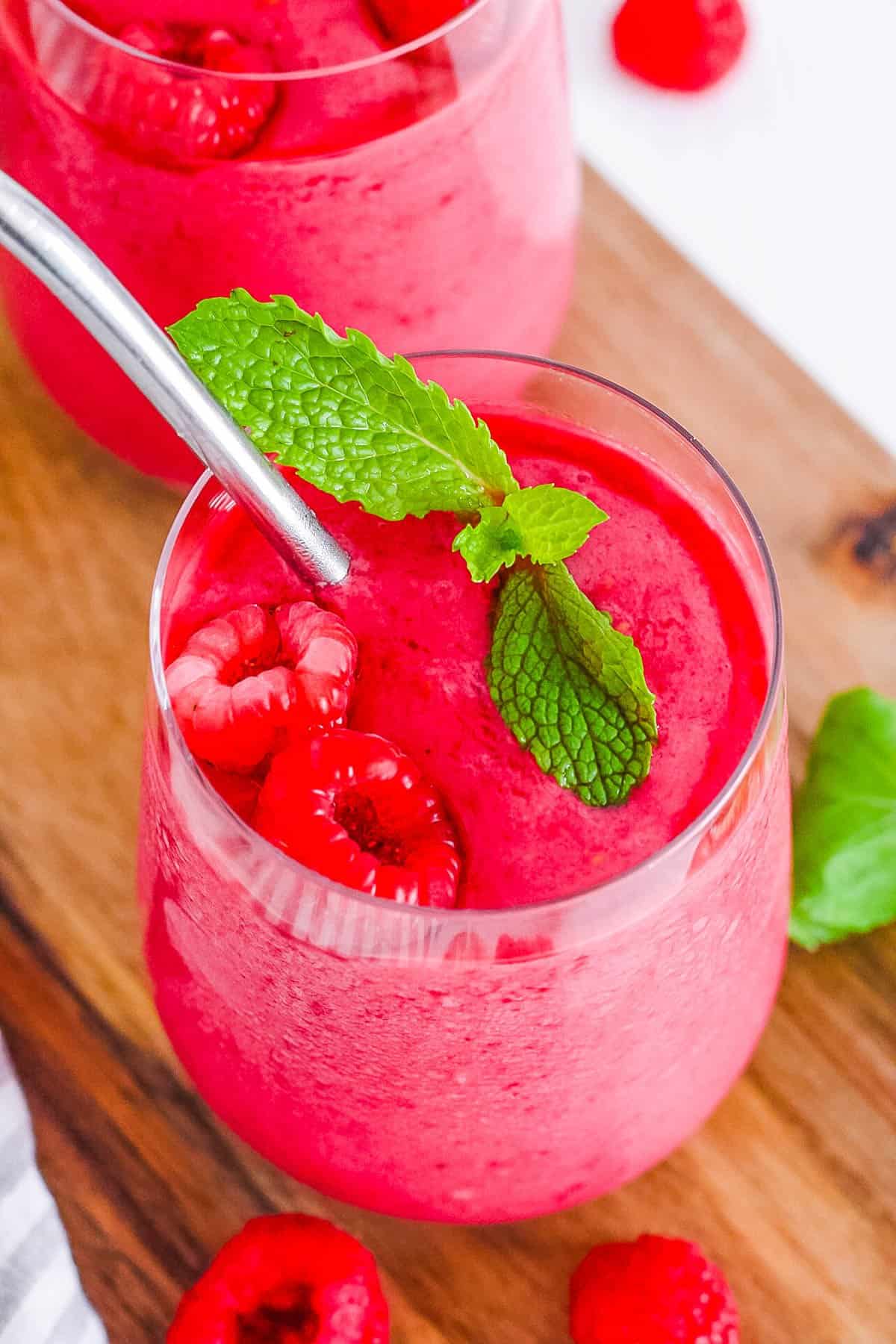 An angled overhead shot of two glasses of raspberry smoothie on a wooden cutting board.